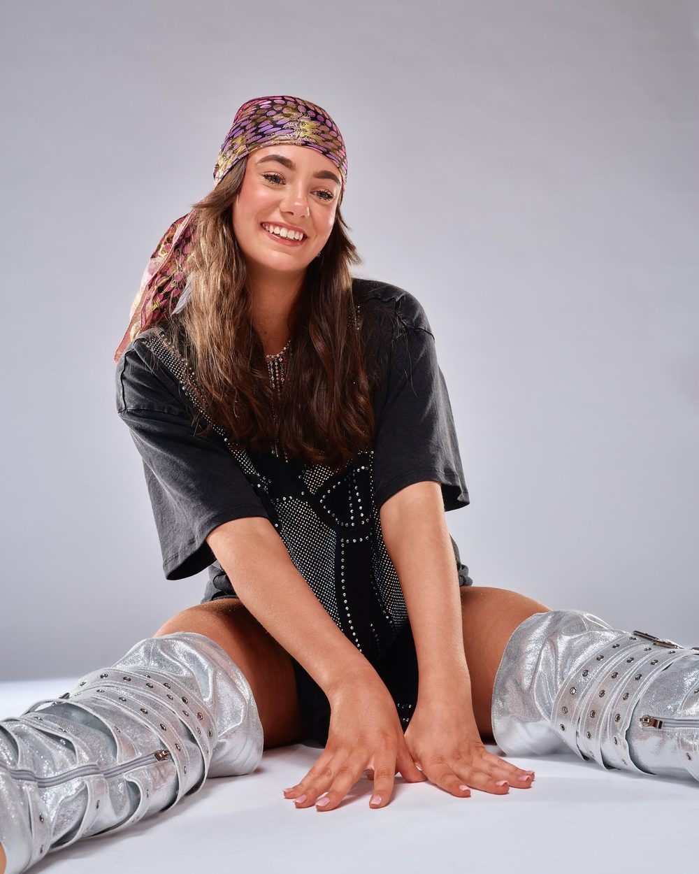 Topaz smiling, sitting on the studio floor in silver boots and a patterned headscarf